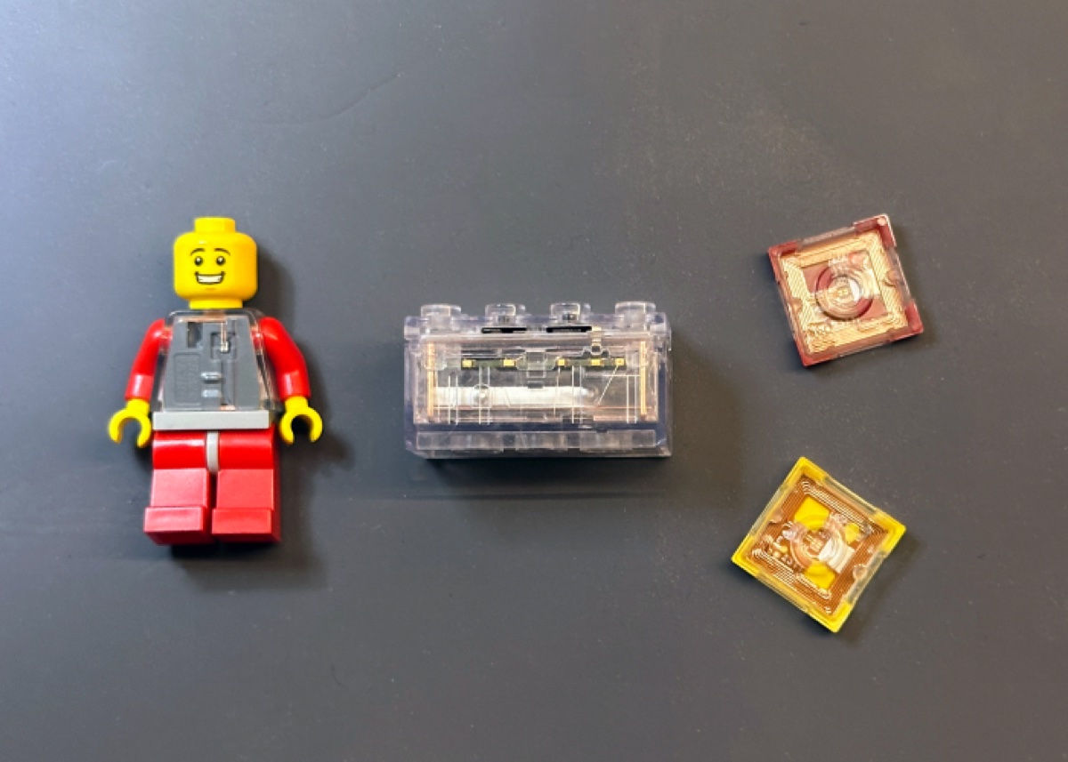 A transparent SMART Brick showing the internal electronics, next to a SMART Minifigure and two SMART Tags